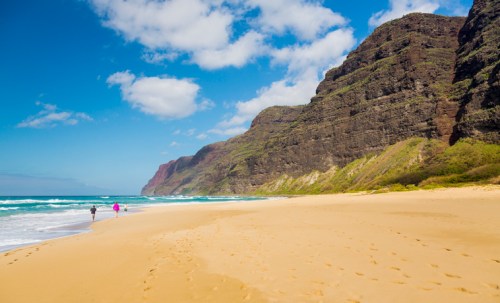 Two people walk along a sandy beach with cliffs and waves under a blue sky with scattered clouds.