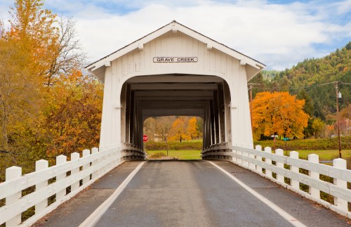 A white covered bridge spans a road, surrounded by autumn trees in vibrant colors under a cloudy sky.