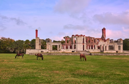 Horses graze in a grassy field near the ruins of a historic building under a cloudy sky.