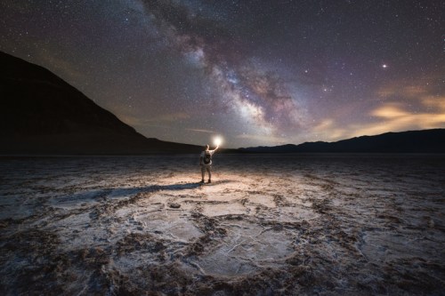 An astronaut stands on a barren landscape under a starry sky, illuminated by the Milky Way and distant mountains.