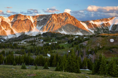 Sunlit mountain peaks rise above a lush green valley, with patches of snow and scattered trees under a blue sky.