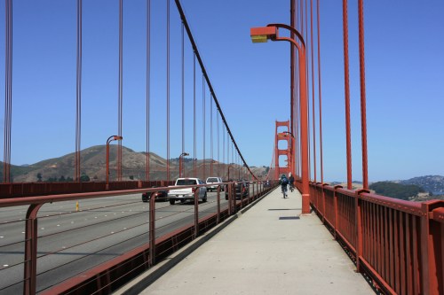 A view of the Golden Gate Bridge with a cyclist and vehicles on the roadway, surrounded by hills and clear blue sky.