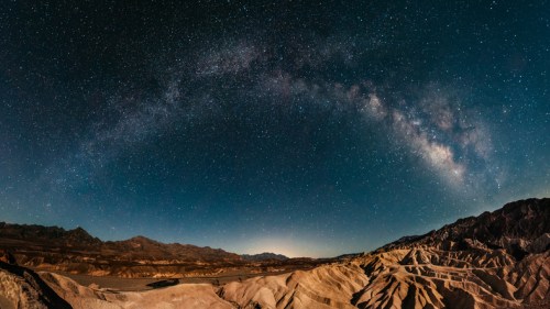 A panoramic view of the Milky Way arching over rugged desert terrain under a starry night sky.