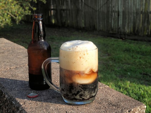 A frothy glass of dark beer sits on a stone surface, with a brown bottle and bottle cap nearby, against a green background.