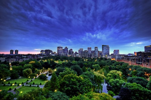 A vibrant city skyline at dusk, surrounded by lush greenery and illuminated park lights under a dramatic sky.