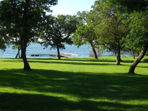 A serene lakeside view with green grass, trees, and a wooden dock extending into the water under a clear blue sky.