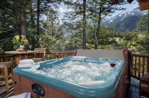 A hot tub on a wooden deck surrounded by trees and mountains, with a glass of wine and flowers nearby.