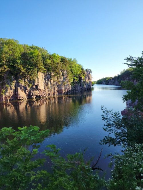 A serene river flows between tall, rocky cliffs, surrounded by lush green trees under a clear blue sky.