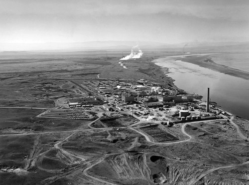 Aerial view of an industrial complex near a river, surrounded by barren land and smoke rising from chimneys.