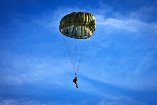 A parachutist descends through a clear blue sky, suspended beneath a green parachute.