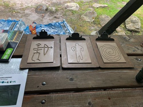 Three wooden boards with drawn shapes: an arrow, a stick figure, and a spiral, placed on a table outdoors.