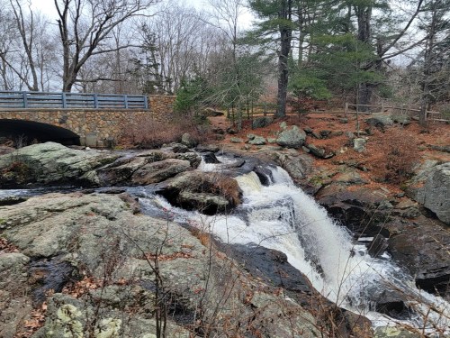 A waterfall cascades over rocks near a stone bridge, surrounded by bare trees and autumn foliage.