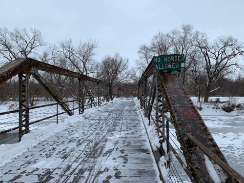A snow-covered metal bridge with a sign reading "No Horses Allowed," surrounded by bare trees and a frozen river.