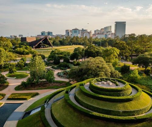 Aerial view of a park with lush greenery, a spiral garden, and a city skyline in the background.