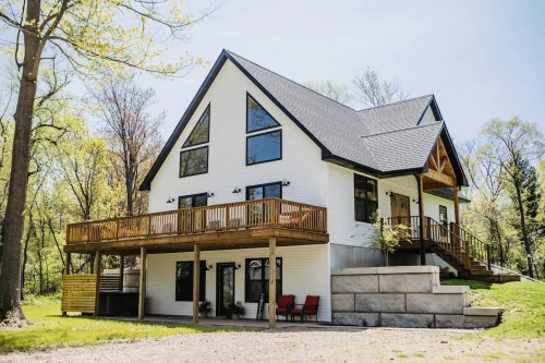 A modern two-story house with a large deck, surrounded by trees and greenery on a sunny day.