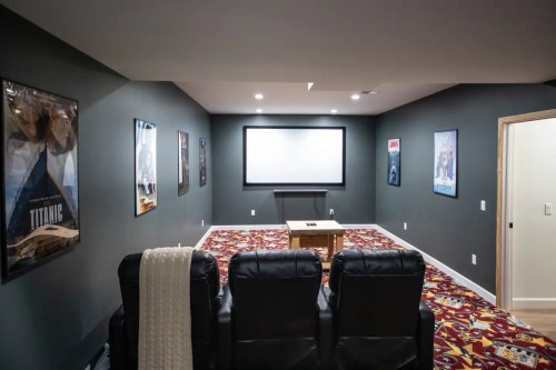 A cozy home theater with black recliners, a large screen, and movie posters on the walls. Colorful carpet adds warmth.