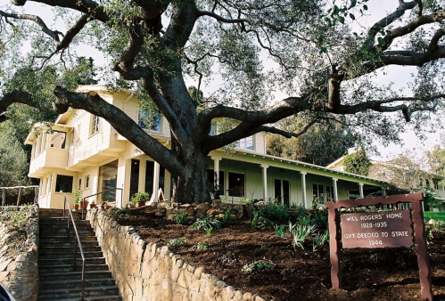Historic Will Rogers Home, featuring a large tree and stone steps, surrounded by greenery and a sign detailing its history.