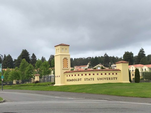 Sign for Humboldt State University, featuring a decorative entrance with trees and buildings in the background under a cloudy sky.