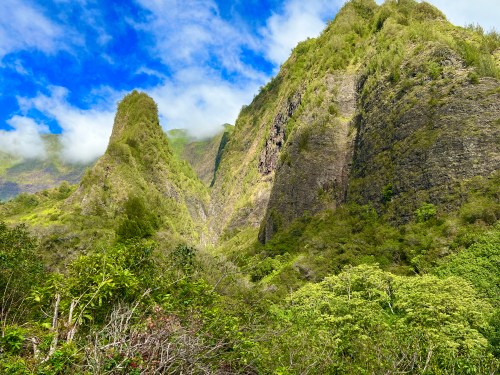Lush green mountains rise under a blue sky with scattered clouds, showcasing a vibrant natural landscape.