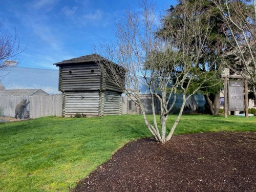 A wooden structure resembling a fort, surrounded by grass and trees, under a clear blue sky.