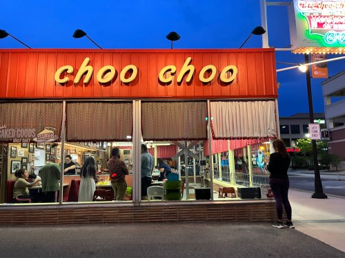 A diner named "Choo Choo" with a warm glow, people dining inside, and a woman standing outside at dusk.