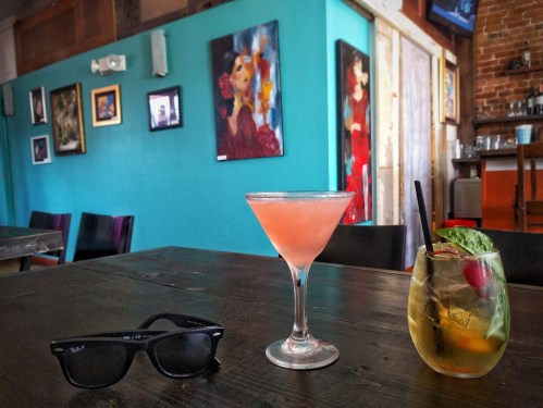 A table with two colorful cocktails and sunglasses, set against a vibrant blue wall with artwork in a cozy bar.