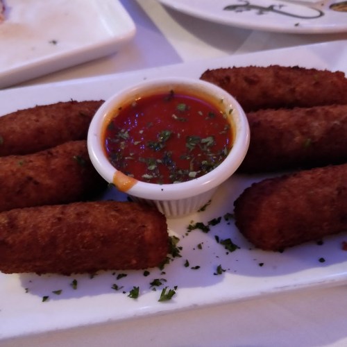 A plate of golden-brown croquettes served with a small bowl of marinara sauce and sprinkled with herbs.