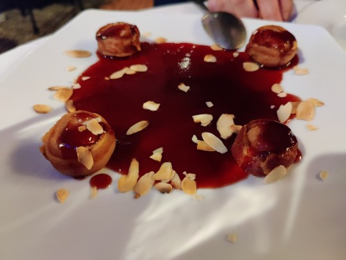 A white plate with three small desserts surrounded by a red sauce and sprinkled with almond slivers.