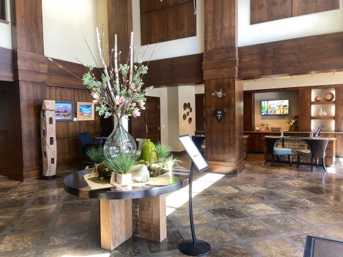 A spacious hotel lobby featuring a floral arrangement, wooden decor, and a reception area with seating.