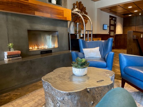 Cozy lounge area with blue chairs, a modern fireplace, and a decorative table with a plant. Warm and inviting atmosphere.