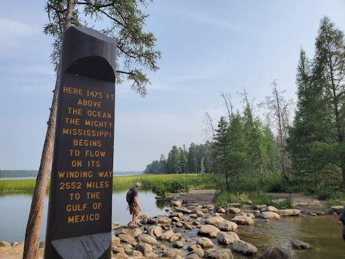 A stone marker by a river notes the Mississippi's origin, with a person walking on rocks nearby and trees in the background.