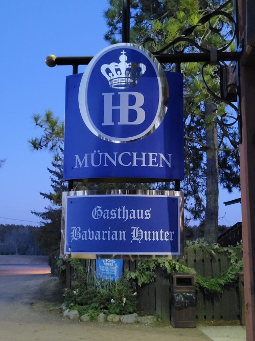 Sign for "Gasthaus Bavarian Hunter" in München, featuring a crown logo, illuminated at dusk.