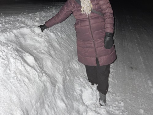 A person in a winter coat stands beside a large snowbank at night, with snow covering the ground.