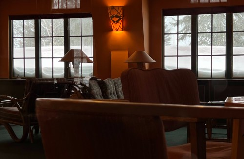 Cozy interior of a room with wooden furniture, lamps, and large windows showing snow outside.