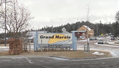 Sign for Grand Marais, featuring directions to the Town Center and Artist Point Boat Launch, with a snowy landscape.