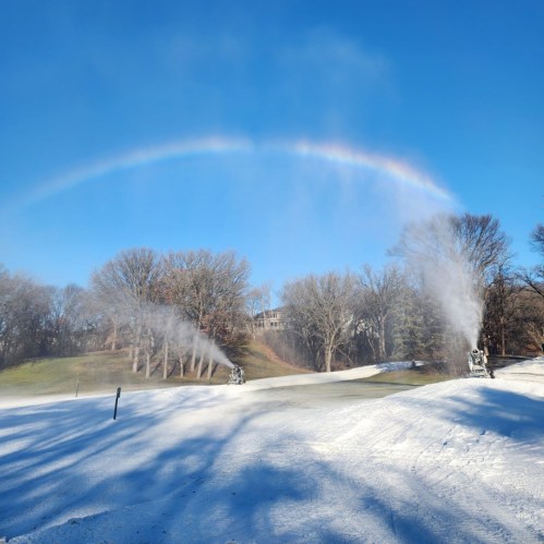 Snowmaking machines operate on a snowy landscape under a clear blue sky, with a rainbow arcing in the background.