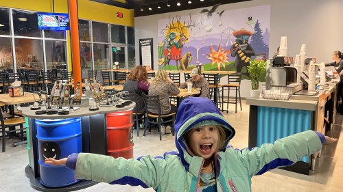 A joyful child in a colorful jacket stands with arms outstretched in a lively restaurant with a mural and patrons in the background.