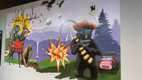 A colorful mural featuring a bear with a gun, animals, a ferris wheel, and whimsical elements like flying cats and food.
