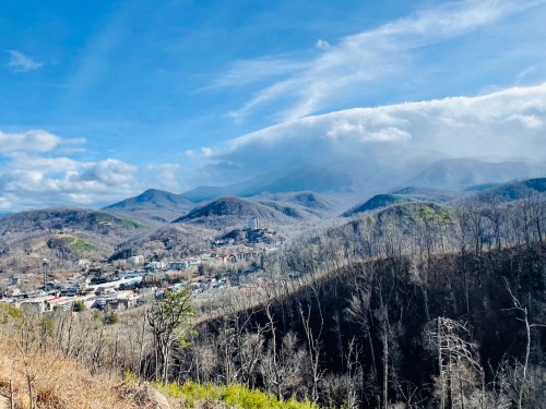 A scenic view of mountains under a blue sky with clouds, overlooking a small town surrounded by bare trees.