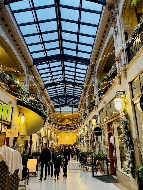 A beautifully decorated indoor shopping arcade with a glass roof, featuring festive lights and people walking.