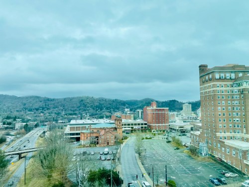 A cloudy view of a cityscape with buildings, mountains in the background, and a mostly empty parking lot.