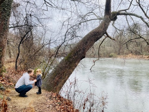 A parent kneels by a riverbank, interacting with a young child near a leaning tree on a cloudy day.