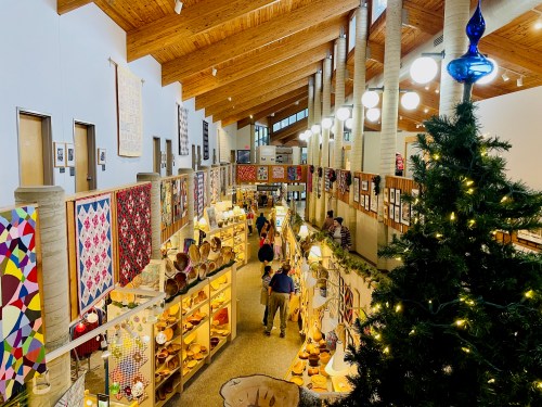 A spacious gallery with wooden beams, showcasing crafts and quilts, decorated for the holidays with a Christmas tree.