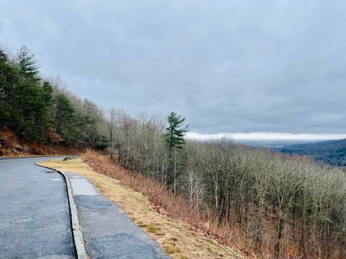 A winding road alongside a forested hillside, with a cloudy sky and a view of a valley below.