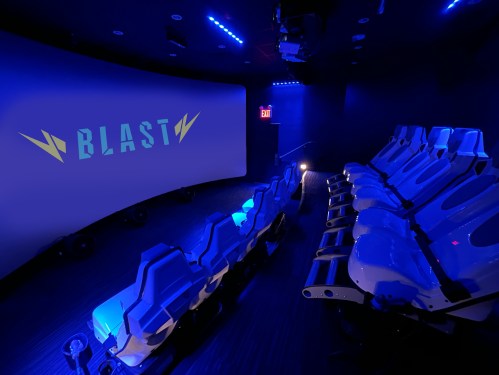 A futuristic theater with blue lighting, featuring rows of white seats and a large curved screen displaying "BLAST."