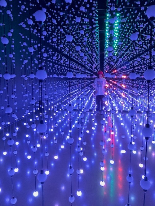 A person stands in a dark room filled with colorful hanging lights, creating a mesmerizing, immersive atmosphere.