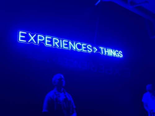 A blue neon sign reads "EXPERIENCES > THINGS," reflecting on a dark background with two people below.