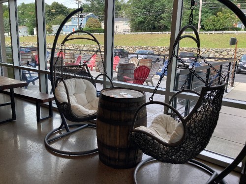 Two hanging chairs with white cushions beside a barrel table, set in a bright, modern indoor space with a view outside.