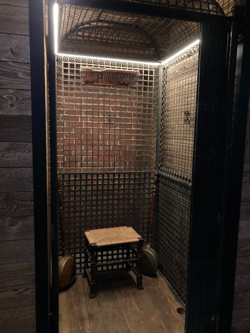 A dimly lit, vintage wooden room with a metal grid, featuring a small stool and a sign reading "The Hoosgow."
