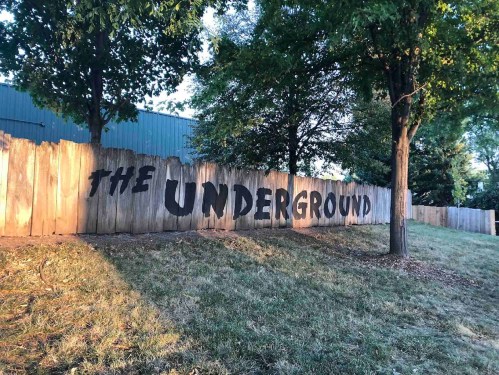 A wooden fence with the words "THE UNDERGROUND" painted in bold black letters, surrounded by trees and grass.